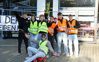 Abiturientinnen und Abiturienten stehen mit Warnwesten bekleidet vor dem Gymnasium