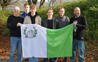 Der Präsident, die Vizepräsidentin, der Oppositionsführer sowie Lehrer aus dem Organisationsteam, stellen die Flagge von Trimon vor.